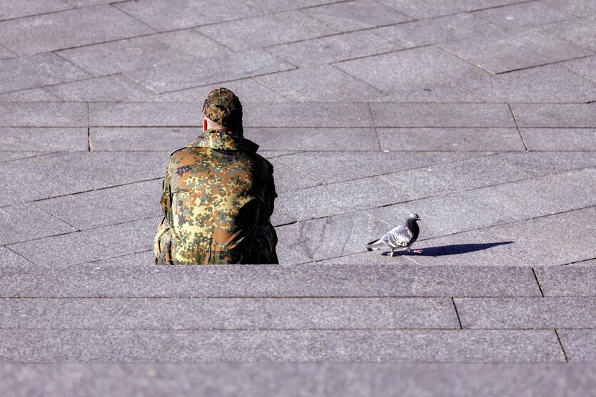 Ein Bundeswehrsoldat sitzt auf einer Treppe, er ist von hinten zusehen, neben ihm, auf dem Straßenpflaster stolziert eine Taube