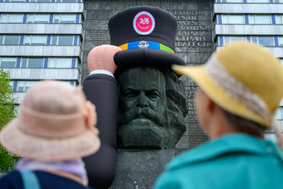 Karl-Marx-Monument in Chemnitz trägt einen aufblasbaren Hut, davor zwei Passant*innen mit Hut