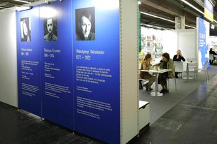 Blick in eine Koje auf der Frankfurter Buchmesse, an einem Tisch sitzen zwei Menschen, an der Wand vor der Koje sind drei große Banner befestigt, mit der Biographie von im Krieg getöteten Autor*innen