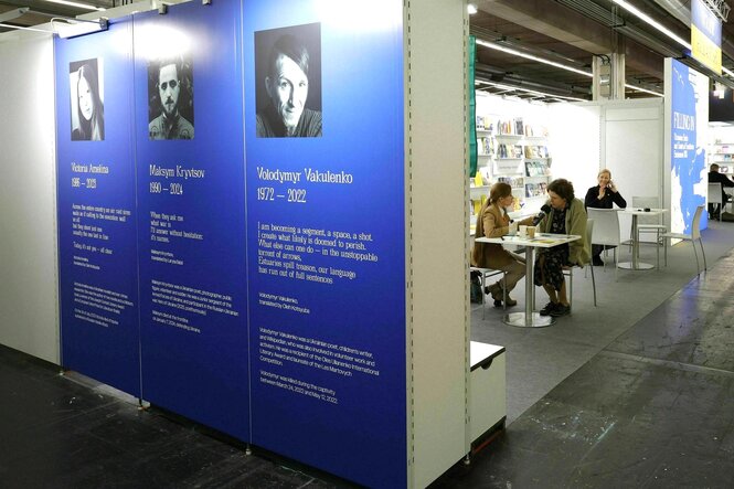 Blick in eine Koje auf der Frankfurter Buchmesse, an einem Tisch sitzen zwei Menschen, an der Wand vor der Koje sind drei große Banner befestigt, mit der Biographie von im Krieg getöteten Autor*innen