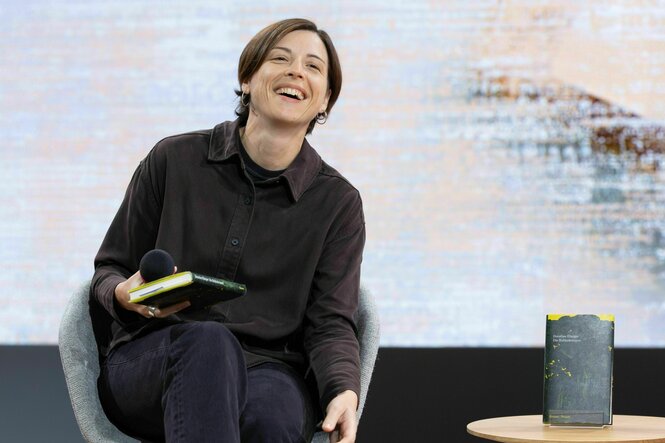 Dorothee Elmiger lacht während eines Interviews auf der Literaturbühne, sie hält ihr Buch in der Hand, ein weiteres Exemplar steht auf einem kleinen tisch neben ihrem Sessel