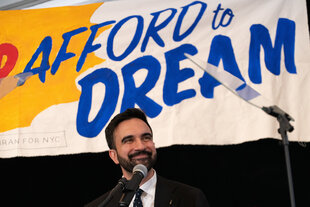 Zoran Mamdani steht auf einer Bühne mit Mikrofon vor einem Banner mit der Aufschrift "Afford to dream on".