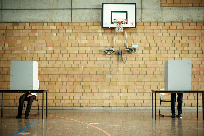 Zwei Personen sitzen hinter ihrer Wahlkabine in der Turnhalle einer Schule in Duisburg