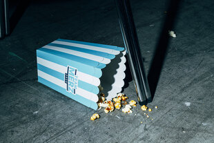 Eine halbleere Pappschachtel Popcorn liegt neben einem Stuhlbein auf dem Boden