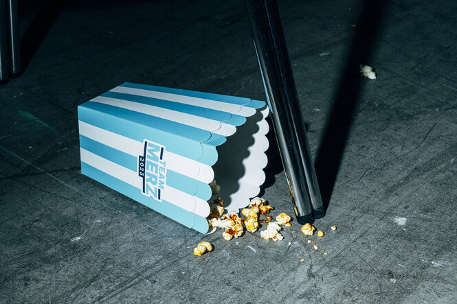 Eine halbleere Pappschachtel Popcorn liegt neben einem Stuhlbein auf dem Boden