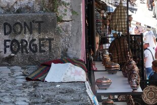 Auf der linken Seite ist eine Mauer zu sehen, mit der Aufschrift „Don't forget“. Auf der rechten Seite sind Waren auf Tischen aufgebaut und es laufen Tourist:innen durch eine Straße.