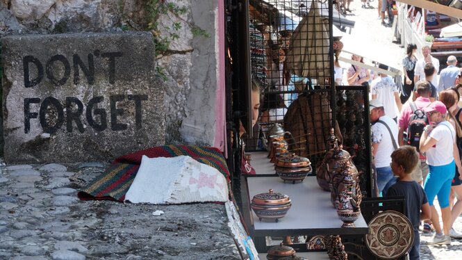 Auf der linken Seite ist eine Mauer zu sehen, mit der Aufschrift „Don't forget“. Auf der rechten Seite sind Waren auf Tischen aufgebaut und es laufen Tourist:innen durch eine Straße.