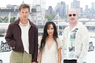 Drei Personen – Austin Butler, Zoe Kravitz und Darren Aronofsky – stehen nebeneinander vor der Stadtkulisse Londons mit hohen, modernen Gebäuden und einem Riesenrad im Hintergrund. Sie blicken in die Kamera und tragen unterschiedliche Outfits in hellen und gedeckten Farben.