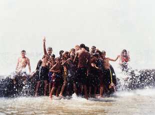 Eine Gruppe spielender Jugendlicher mit Badeshorts und Badeanzug bilden ein Knäuel im trüben Brackwasser an einem warmen Strand, spritzen und toben