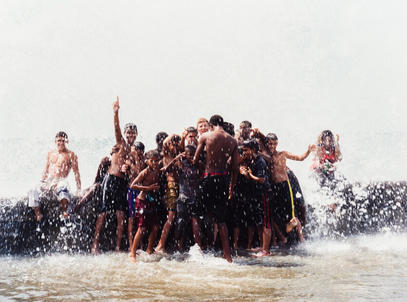 Eine Gruppe spielender Jugendlicher mit Badeshorts und Badeanzug bilden ein Knäuel im trüben Brackwasser an einem warmen Strand, spritzen und toben