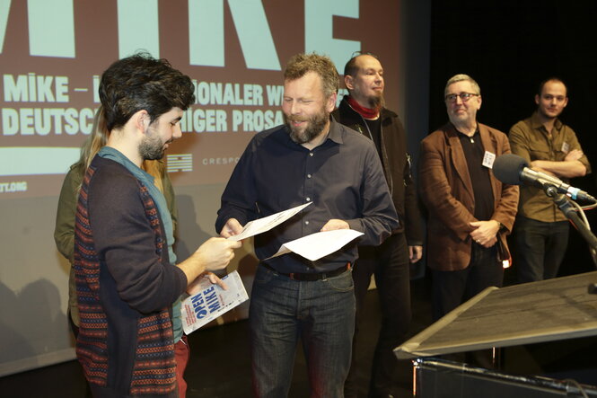 Auf dem Foto sind der Presiträger des open mikes 2024 Gerasimos Bekas und der Ressortleiter Kultur der taz Andreas Knipphals zu sehen, der Gerasimos Bekas die Urkunde überreicht.