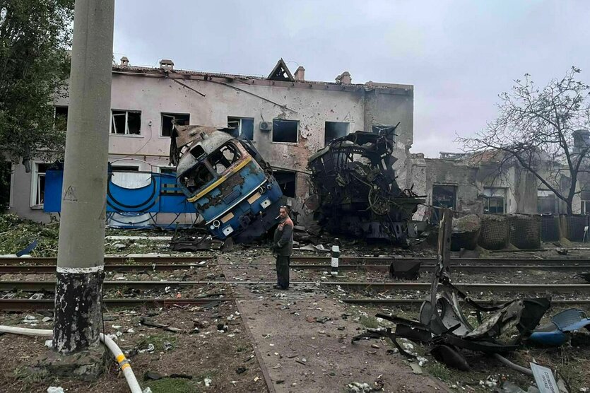 EIn zerstörter Bahnübergang, eine Lok und neben ihr ein ausgebrannter Wagen liegen neben den Gleisen, eine verdreckte Person blickt uns über die Schulter an.