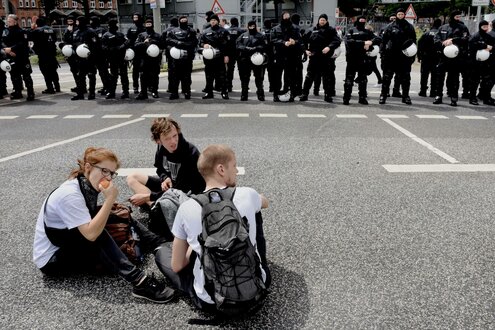 drei Menschen sitzen auf der Straße vor einer Reihe Polizisten