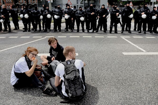 drei Menschen sitzen auf der Straße vor einer Reihe Polizisten