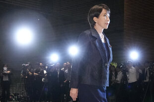 Sanae Takaichi verlässt ein Gebäude, sie trägt schwarz, es ist Nacht und im Hintergrund ist ein Reihe Journalist*innen zu sehen