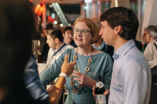 Menschen unterhalten sich in entspannter Atmosphäre bei einer Veranstaltung, eine Frau mit Brille hält ein Glas Sekt, ein Mann daneben eine Flasche Bier.