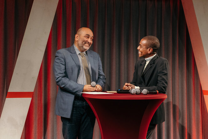 Omid Nouripour steht neben der Moderation auf der Bühne der taz Kantine, beide lächeln.