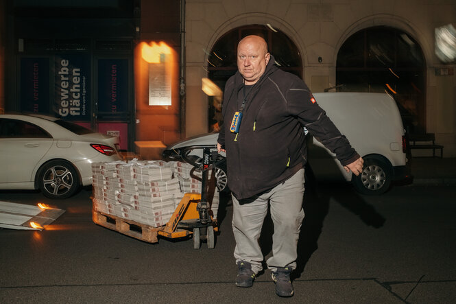 Ein Mann lehnt sich mit voller Kraft in die Rollrichtung seines Hubwagens, der voll mit Zeitungen gestapelt ist.