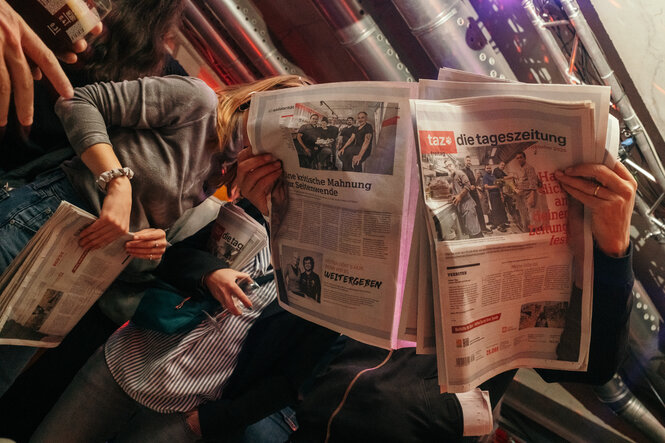 Auf dem Foto sind mehrere Personen zu sehen, die in Zeitungen lesen. Die Zeitungen tragen den Titel „taz – die tageszeitung“.