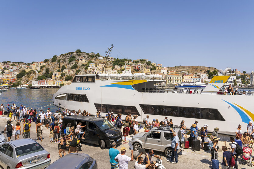 Passagiere tummeln sich, sie warten zum Einstieg auf ein Fährboot am Hafen von Symi mit der farbenfrohen, griechischen neoklassizistischen Architektur, im Hafen liegen Segelboote, große Luxusmotoryachten und einfache Fischerboote. Auf dem Parkplatz vor der Fähre stehen mehrere Autos. Der Himmel ist strahlend blau.