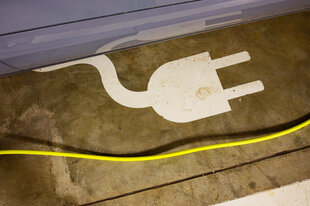 Auf einem Parkplatz ist das Symbol eines Ladesteckers an einem Kabel markiert. Ein gelbes Ladekabel hängt knapp über dem Boden. Ein Teil der Markierung auf dem Parkplatz wird von einem Auto verdeckt, ein kleiner Teil der glänzenden Karosserie ist mit auf dem Foto.
