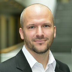 Gregor Kolbe ist eine weiße Person mit hellbraunem Dreitagebart. Er trägt ein weißes Hemd und ein schwarzes Sakko und lächelt mit geschlossenem Mund in die Kamera.