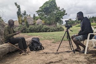Zwei Männer sitzen auf Baumstämmen, in ihrer Mitte eine Kamera