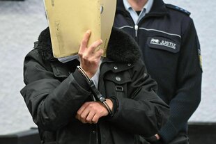 Ein Mann hält sich einen Aktendeckel vor das Gesicht und wird in Handschellen in den Gerichtssaal geführt