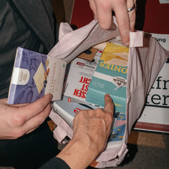 Goodiebag der taz Seitenwende: Stofftasche mit Schokolade und anderen Esswaren sowie Printprodukten. Hände greifen in die Tasche hinein und holen sich Artikel raus.