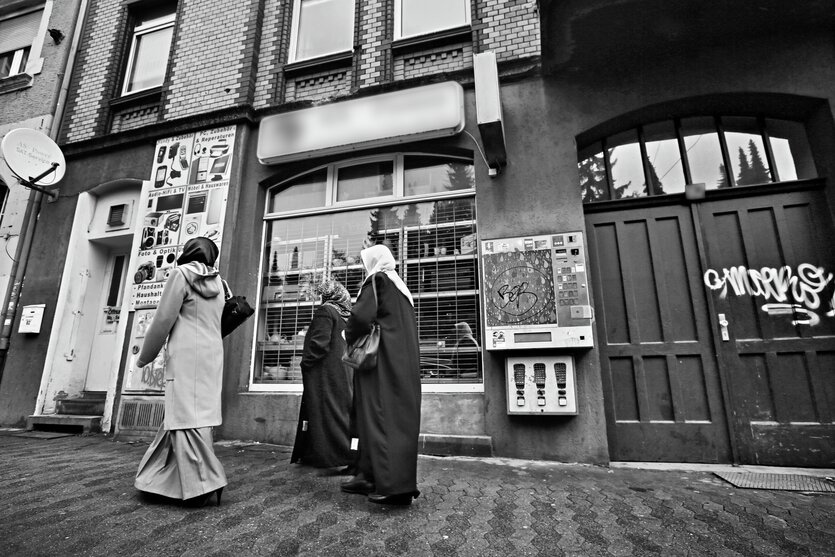 Ein Schwarz-Weiß-Foto eines Geschäfts, vor dem drei Frauen mit Kopftüchern auf dem Bürgersteig laufen
