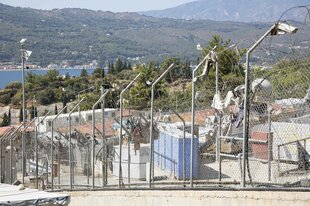 Blick auf eine Bucht, halbversperrt von einen Zaun mit einer Überwachungskamera, dahinter ine Lager für Geflüchtete