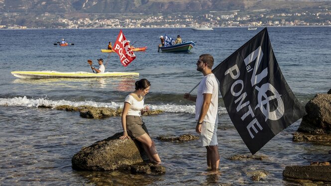 Menschen protestieren am Strand, eine trägt eine schwarze Fahne mit der weißen Aufschrift 