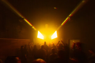 „Menschen tanzen in einem dunklen Nachtclub in Kyiv, beleuchtet von intensivem gelbem Licht, das aus rechteckigen Scheinwerfern auf der Bühne strahlt. Die Szene ist verschwommen und vermittelt eine energiegeladene, atmosphärische Stimmung.“