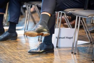 Ein paar Beine in Lederschuhen, die nicht erkennbare Person sitzt in einer Stuhlreihe. Auf einem Papierzettel zwischen den Stuhlbeinen steht der Name: Manuel Hagel.
