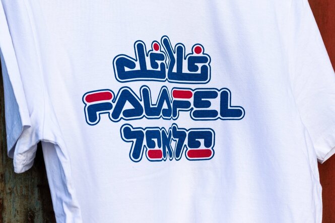 Ein weißes T-Shirt, auf dem das Wort Falafel in arabischer, lateinischer und hebräischer Schrift steht