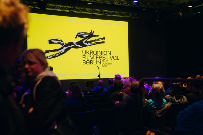 Menschen sitzen in einem abgedunkelten Saal und warten auf den Beginn einer Veranstaltung. Auf einer großen gelben Leinwand steht „Ukrainian Film Festival Berlin, 22–26 October 2025“ mit einer schwarzen Grafik eines stilisierten laufenden Pferdes.