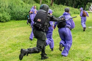 Ein Polizist der Beweissicherungs- und Festnahmeeinheit läuft mit gezogenem Schlagstock einer Gruppe von Aktivist:innen in Lila Overalls auf einer Wiese hinterher.