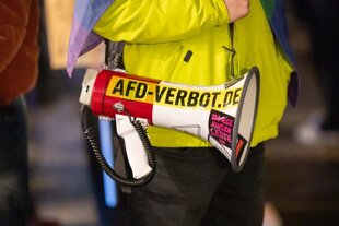 Foto eines Megaphones, das ein Demonstrant in Gelber Jacke geschultert hat. Darauf wurde mit schwarzen Lettern auf gelben Grund „Afd-Verbot.de“ geklebt.