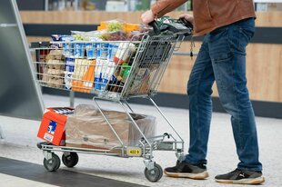 Ein Mensch, von dem nur der Unterkörper zu sehen ist, schiebt einen vollen Einkaufswagen