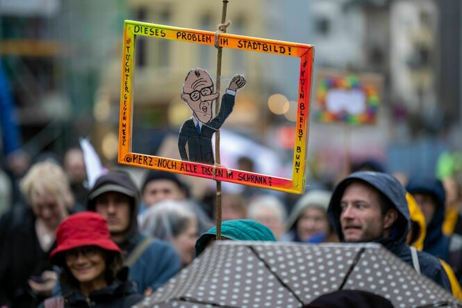 Eine Teilnehmerin hält eine aus Pappe ausgeschnittene Figur von Kanzler Merz hoch