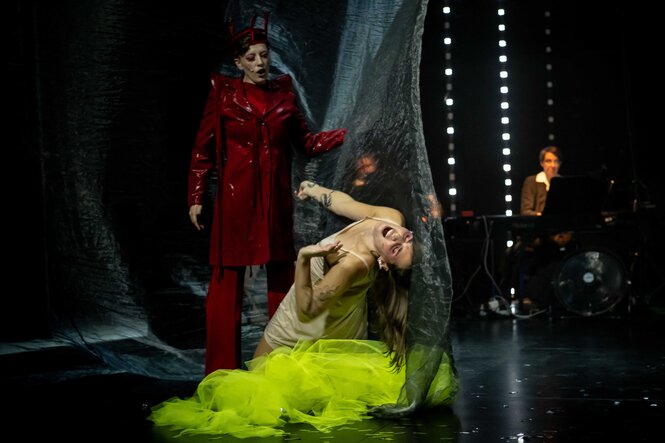 Eine Figur in rotem Mantel steht vor einer tätowierten Frau in weißem Slipkleid und gelbem Tüllrock, die sich schreiend und verdreht in durchsichtigem Vorhang windet. Im Hintergrund ein Pianist bei gedimmtem Licht, Trommeln und Ventilator.
