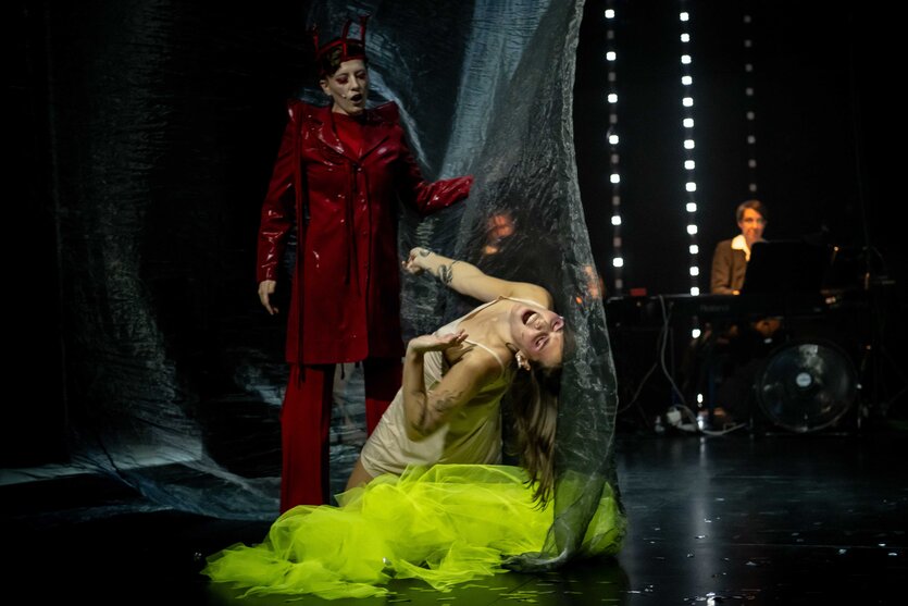 Eine Figur in rotem Mantel steht vor einer tätowierten Frau in weißem Slipkleid und gelbem Tüllrock, die sich schreiend und verdreht in durchsichtigem Vorhang windet. Im Hintergrund ein Pianist bei gedimmtem Licht, Trommeln und Ventilator.