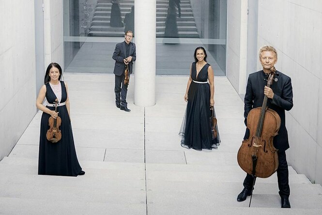 Auf dem Bild sind vier Musiker:innen des Minguet Quartetts zu sehen, die teils auf einer hellgrauen Treppe vor einer Glaswand stehen und Streichinstrumente in der Hand halten.