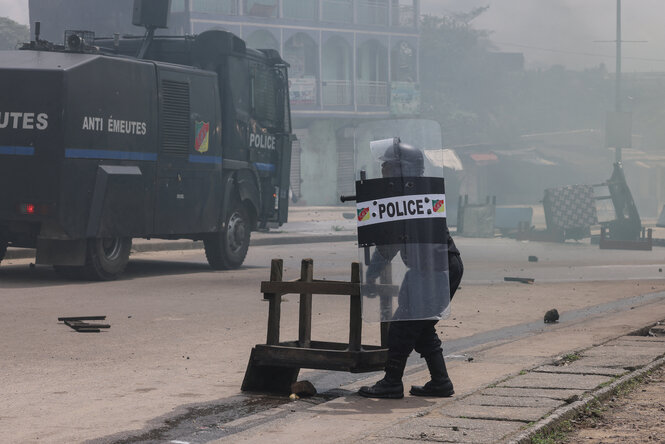 Polizist duckt sich hinter einem Schild, im Hintergrund fährt ein Wasserwerfer