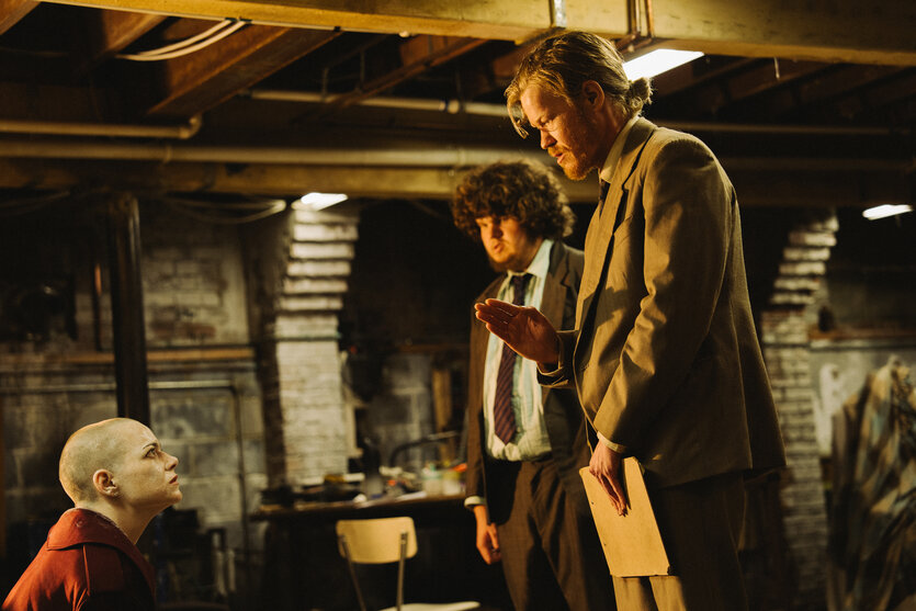 Drei Personen in einem Kellerraum: Eine kahlrasierte Frau in rotem Hemd kniet und blickt auf, zwei Männer in Anzügen stehen daneben, einer hält eine Mappe und hebt die Hand.