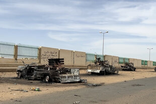 Ausgebrannte Militär-Trucks stehen vor einer mit arabischen Lettern besprayten Mauer.