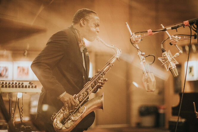 Mann in dunklem Anzug spielt Saxophon, Mikrofone davor, warmer Sepia-Ton.