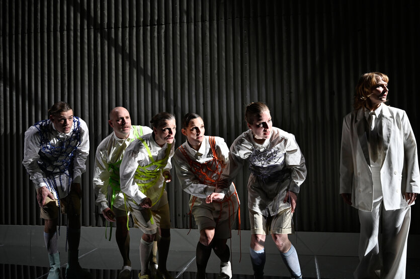 Fünf Personen in weißen Hemden und Shorts, mit bunten Seilen umwickelt, stehen gebückt auf Bühne; eine Frau in weißem Anzug steht daneben.