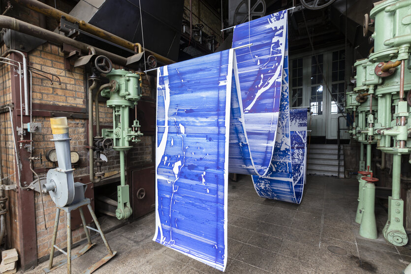 In einem Maschinenraum läuft eine blaue Papierbahn über verschiedene Stangen