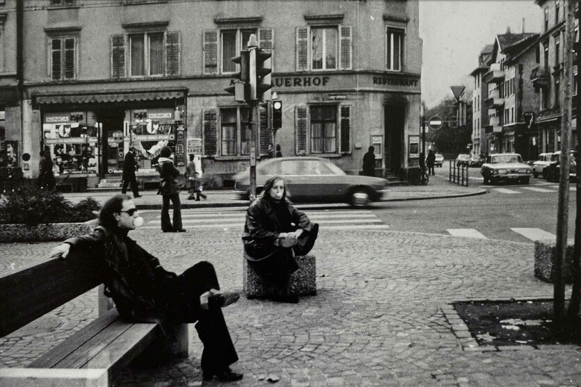 schwarz-weiss Fotografie von zwei personen auf einem Platz, mit Kaugummi-Blase im Mund